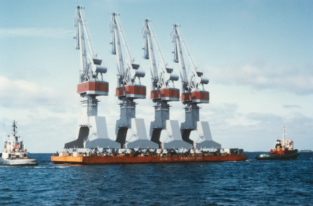 Kaksi hinausvenettä vetää Koneen proomua, jossa seisoo neljä identtistä satamanosturia. Två bogserbåtar drar Kones pråm på vilken det står fyra identiska hamnlyftkranar. Two tugboat transporting a barge with four identical harbor cranes.